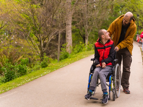 Paralyzed young male in a wheelchair being pushed by a friend in a sunny park